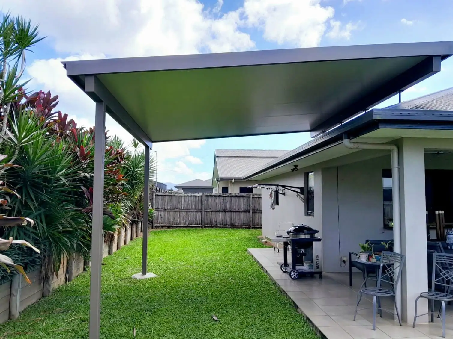 Flat-roof patio cover over a paved outdoor living area, with lawn, planted garden edge and timber fencing